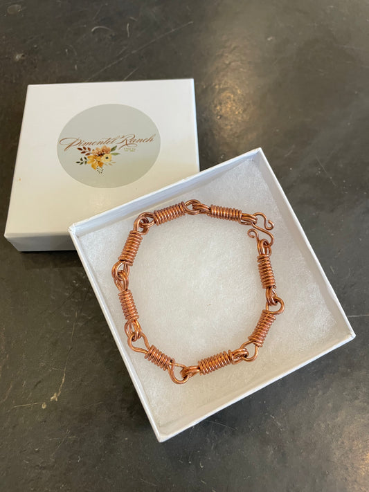 A bracelet made out of hand twisted copper wire with an S hook closure, presented in a white box with a brand label.