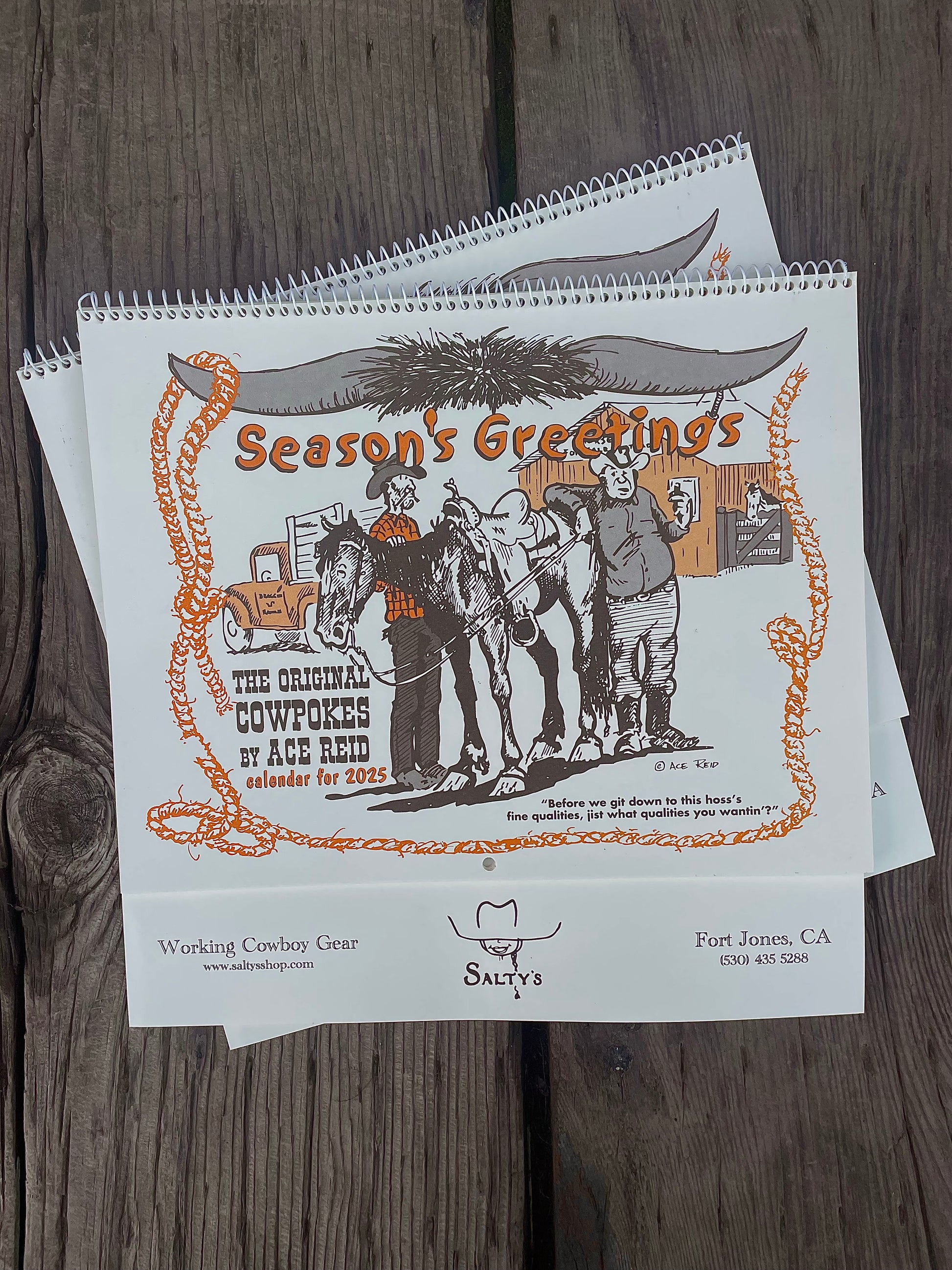 Stack of 'Season's Greetings' Ace Reid Cowpokes calendars on a wooden surface.